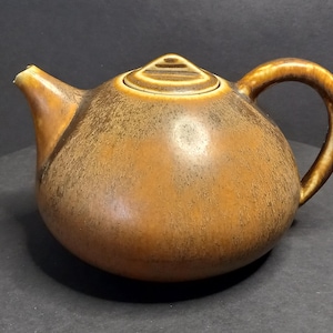 Op de afbeelding: Een bruine keramische theepot met een afgeronde body, een gebogen handvat en een tuit. Het deksel heeft een kleine, gegroefde knop. De theepot heeft een gespikkelde, getextureerde oppervlakte en een glanzende afwerking.