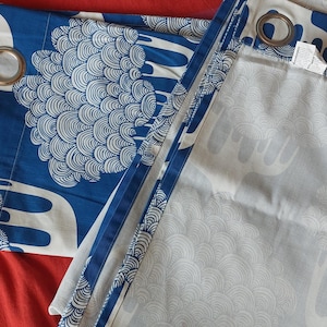 Puede incluir: Cortinas estampadas en azul y blanco con ojales met&aacute;licos. Las cortinas presentan un dise&ntilde;o repetitivo de formas estilizadas. La tela est&aacute; doblada sobre una superficie a rayas rojas y rosas.