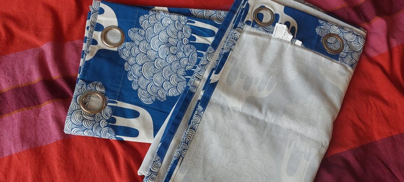 Puede incluir: Cortinas estampadas en azul y blanco con ojales met&aacute;licos. Las cortinas presentan un dise&ntilde;o repetitivo de formas estilizadas. La tela est&aacute; doblada sobre una superficie a rayas rojas y rosas.