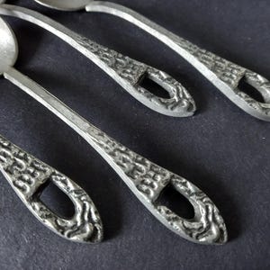 May include: Close-up of several silver-colored spoons. The spoons have textured handles with a decorative pattern and a small opening. The spoons are arranged diagonally against a dark background, showcasing their design.
