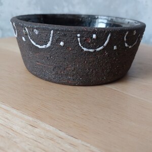 May include: A dark brown ceramic bowl with a white design around the rim. The design features a series of curved lines and dots.