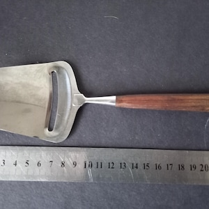 Könnte beinhalten: Ein Käseschneider aus Edelstahl mit Holzgriff. Die rechteckige Klinge hat eine gebogene Kante und einen Schlitz. Der Griff ist dunkelbraun. Ein Lineal liegt neben dem Schneider, mit Messungen in Zentimetern.