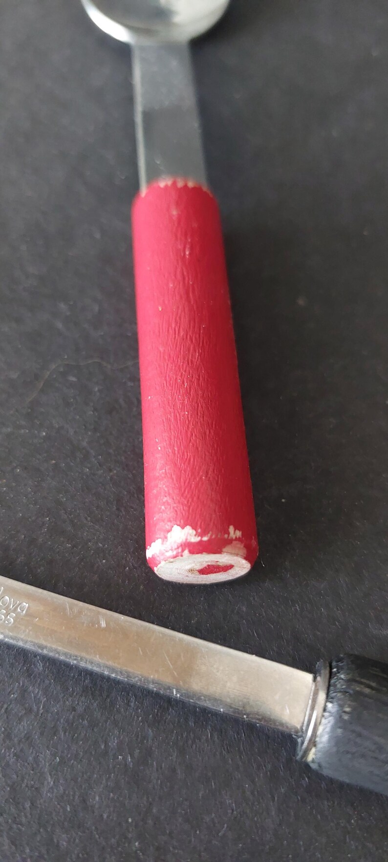 May include: Close-up of a spoon with a red handle and a silver blade. The red paint on the handle is chipped, revealing the wood underneath. Another spoon with a black handle is partially visible.