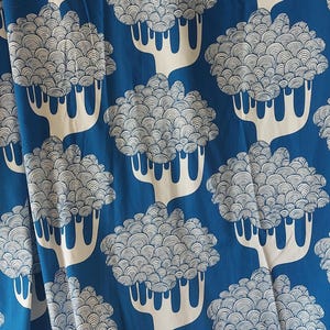 Puede incluir: Cortina estampada en azul y blanco. La tela presenta un dise&ntilde;o repetitivo de &aacute;rboles blancos estilizados con copas redondeadas y en capas sobre un fondo azul liso. La cortina tiene ojales met&aacute;licos para colgar.