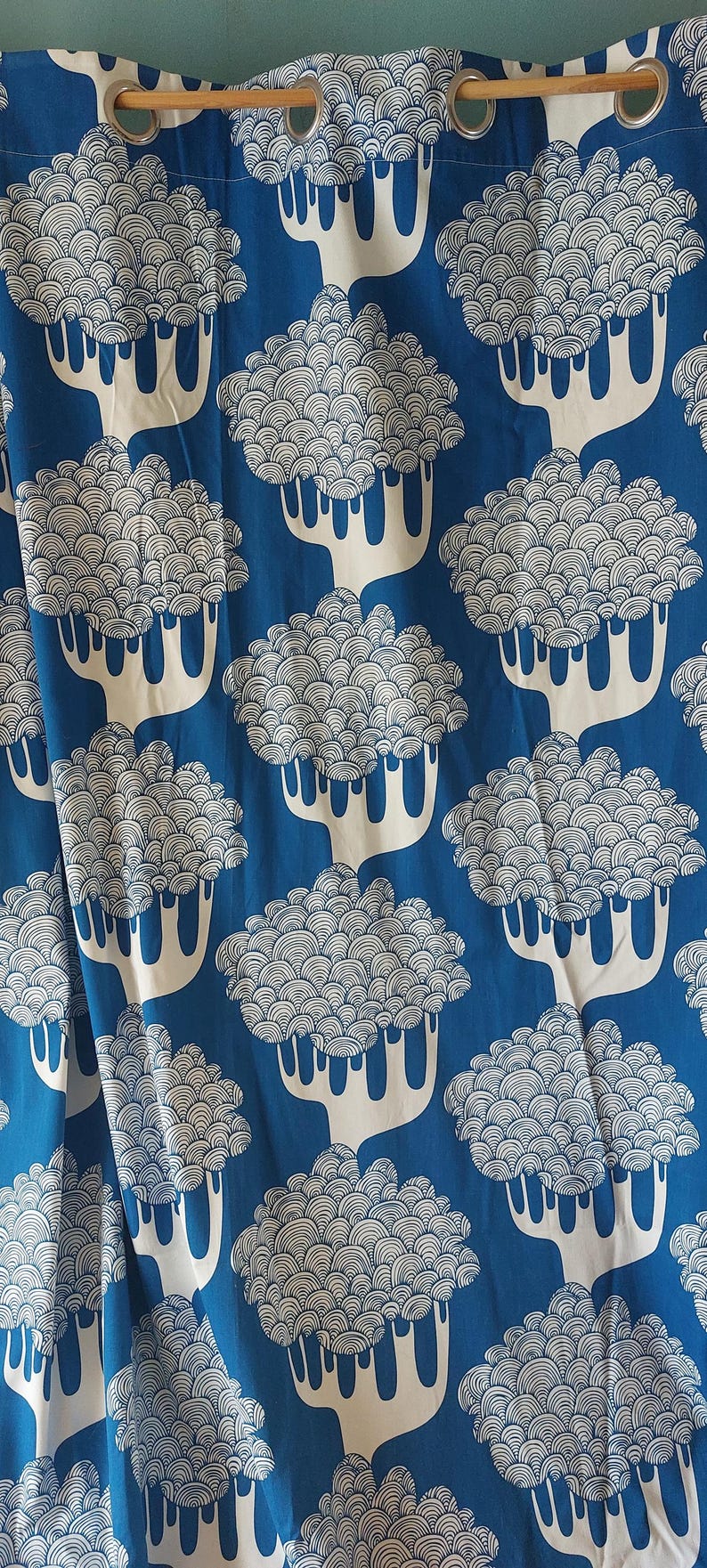 Puede incluir: Cortina estampada en azul y blanco. La tela presenta un dise&ntilde;o repetitivo de &aacute;rboles blancos estilizados con copas redondeadas y en capas sobre un fondo azul liso. La cortina tiene ojales met&aacute;licos para colgar.