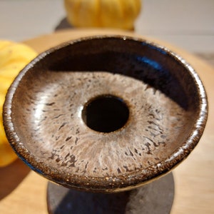 May include: A handmade, brown ceramic candle holder with a unique donut-shaped design. The holder has a dark brown base and a lighter brown interior with a textured surface. The rim is a darker brown. The candle holder is on a wooden surface.