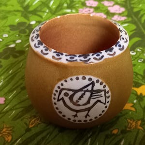 Könnte beinhalten: Eine kleine, braune Keramiktasse mit einem weißen Vogelmotiv. Die Tasse hat einen weißen Rand mit schwarzen und blauen Akzenten.