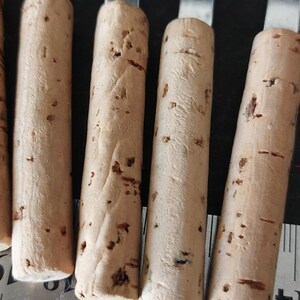May include: Close-up of several cork bottle stoppers with stainless steel handles. The corks are cylindrical, light brown, and have dark brown spots. A ruler is visible in the foreground, indicating the stoppers' size.