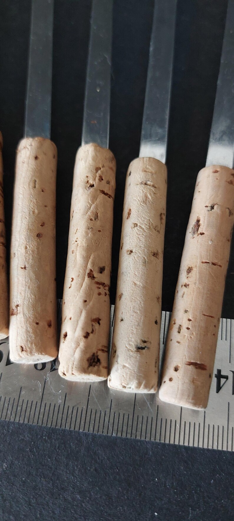 May include: Close-up of several cork bottle stoppers with stainless steel handles. The corks are cylindrical, light brown, and have dark brown spots. A ruler is visible in the foreground, indicating the stoppers' size.