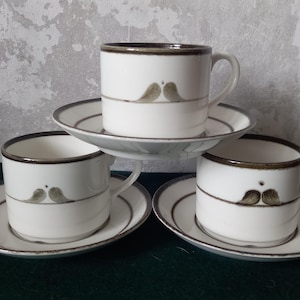 May include: Three white ceramic teacups with brown trim and a stylized bird design. Each cup is on a matching saucer.