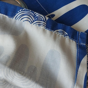 Puede incluir: Primer plano de tela azul y blanca con un patr&oacute;n de olas. La tela es de un azul vibrante con dise&ntilde;os blancos en forma de olas. Los bordes est&aacute;n cosidos cuidadosamente con un borde azul, y se ven ojales met&aacute;licos.