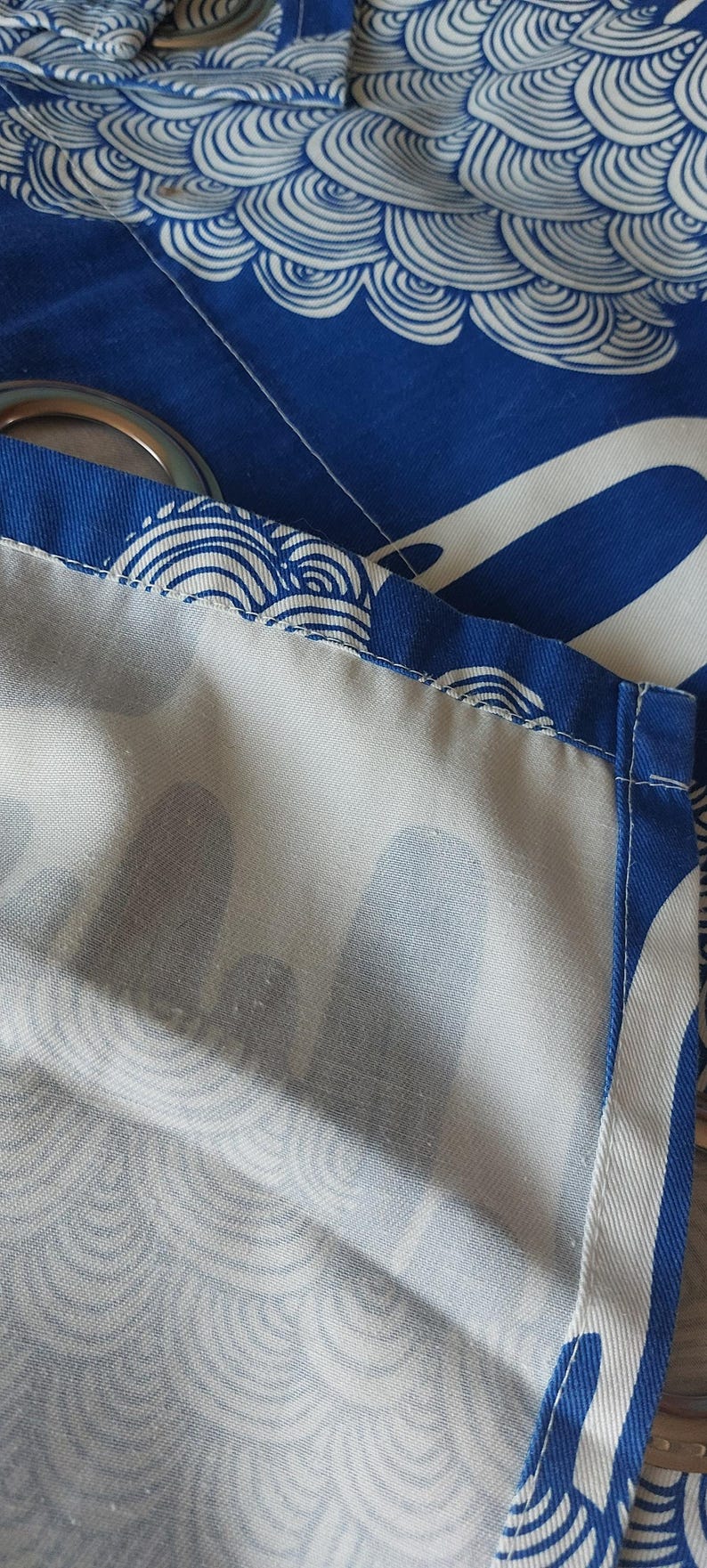 Puede incluir: Primer plano de tela azul y blanca con un patr&oacute;n de olas. La tela es de un azul vibrante con dise&ntilde;os blancos en forma de olas. Los bordes est&aacute;n cosidos cuidadosamente con un borde azul, y se ven ojales met&aacute;licos.