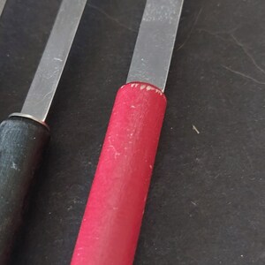 May include: Close-up of several spoons with stainless steel bowls and long, flat handles. One spoon has a red wooden handle, while another has a black wooden handle. The spoons are arranged on a dark surface.