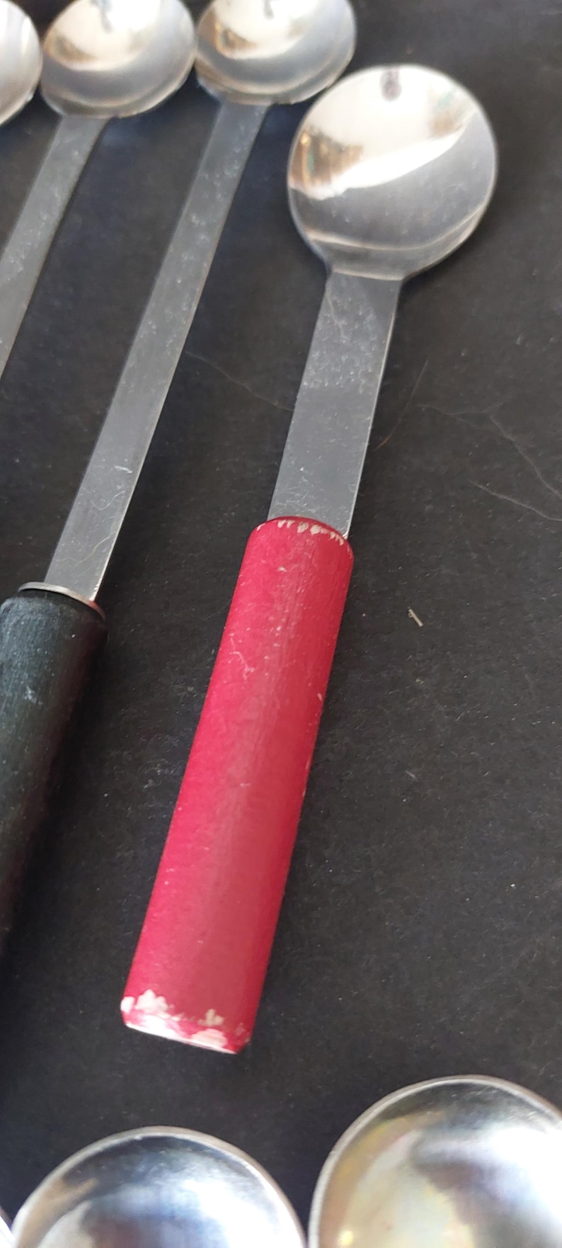 May include: Close-up of several spoons with stainless steel bowls and long, flat handles. One spoon has a red wooden handle, while another has a black wooden handle. The spoons are arranged on a dark surface.