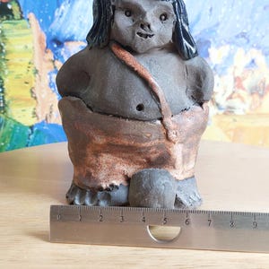 May include: A small ceramic figurine of a person with dark brown skin, black hair, and a brown and orange loincloth. The figurine is about 5 inches tall.