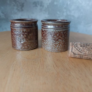 May include: Two small brown ceramic jars with a white glaze and brown lettering. The jars are labeled "HOGAN" and "GANES" with a symbol of an anchor. A cork with a brown and white design is in the foreground.