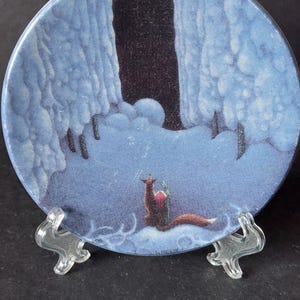May include: A decorative plate with a whimsical illustration of a fox in a snowy landscape under a crescent moon. The plate is supported by a clear plastic stand, and a ruler is in the foreground.