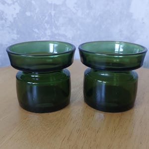 以下が含まれることがあります： 幅広の丸いトップと、より小さい丸いベースを持つ、濃い緑色のガラス製のキャンドルホルダー2つ