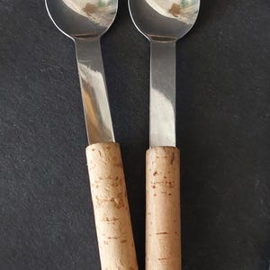 May include: Two stainless steel spoons with round bowls and flat handles. The handles are cylindrical and made of cork. The spoons are on a dark surface.