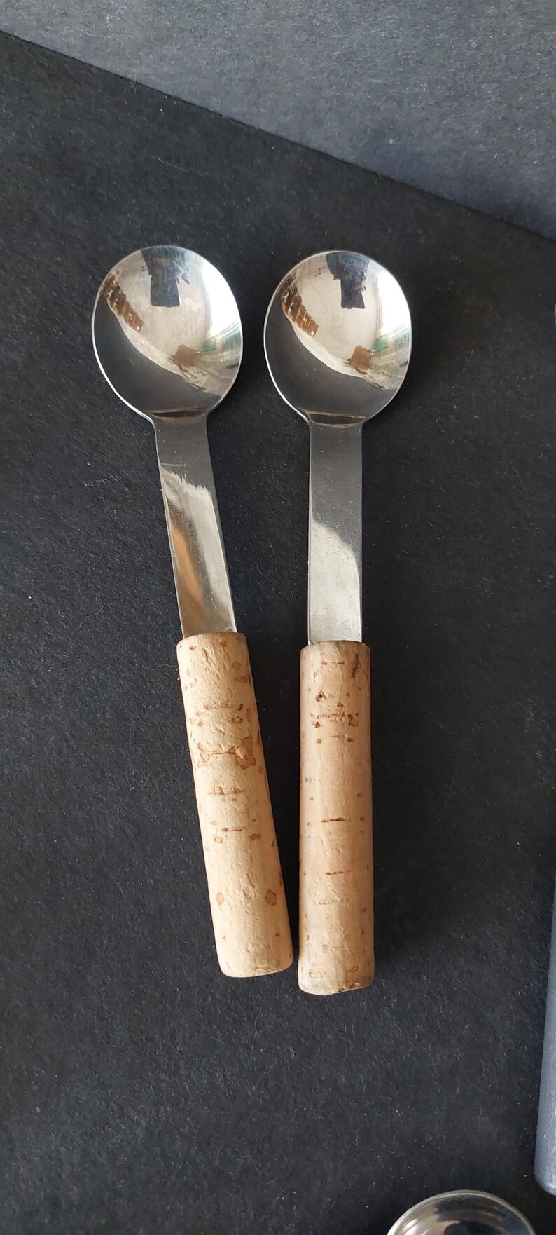 May include: Two stainless steel spoons with round bowls and flat handles. The handles are cylindrical and made of cork. The spoons are on a dark surface.