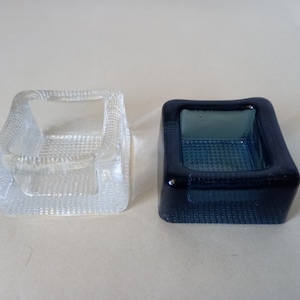 Puede incluir: Dos pequeños platos cuadrados de vidrio, uno transparente y otro azul. Ambos platos tienen una superficie texturizada.