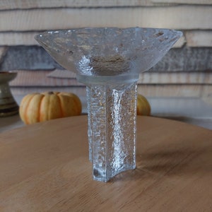 May include: A clear glass candle holder with a textured, frosted finish. The holder has a wide, shallow bowl and a tall, rectangular base.