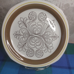 Puede incluir: Un plato de cerámica blanco con un borde marrón y amarillo. El plato presenta un diseño floral marrón simétrico en el centro. El plato se exhibe sobre un soporte metálico sobre un fondo a cuadros azul y verde.