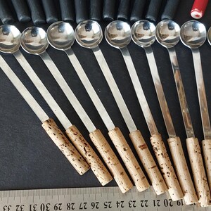 May include: A collection of stainless steel spoons with long, slender handles and cork-like grips. The spoons are arranged in a row, with the rounded bowls facing the viewer. A ruler is visible at the bottom.