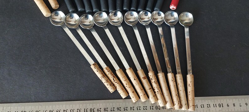 May include: A collection of stainless steel spoons with long, slender handles and cork-like grips. The spoons are arranged in a row, with the rounded bowls facing the viewer. A ruler is visible at the bottom.