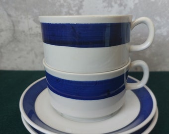 May include: Two white ceramic teacups with blue stripes stacked on top of each other. The cups are on a white saucer with a blue stripe around the edge. The saucer is on top of another white saucer with a blue stripe around the edge.