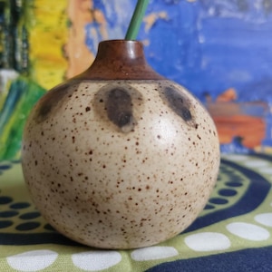 May include: A small, speckled, cream-coloured ceramic vase with brown accents. The vase has a rounded shape and a small opening at the top.