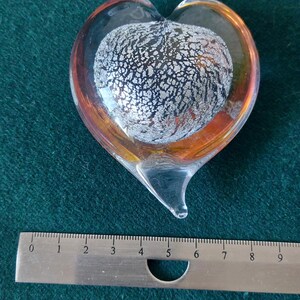 May include: A glass heart-shaped paperweight with a silver and black swirl design inside. The heart is clear glass with a slight orange tint.