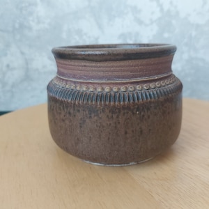 Peut inclure: Un pot en céramique brun avec une surface texturée et une bande décorative autour du haut.