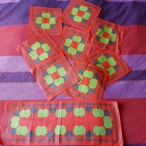 Puede incluir: Conjunto de 10 manteles individuales de tela roja y verde con un patrón de manzanas estilizadas y corazones. Los manteles individuales están dispuestos en forma escalonada sobre un fondo de tela a rayas morado y rojo.