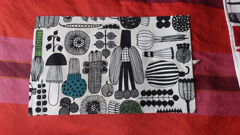 May include: A white kitchen towel with a black and white patterned design featuring various shapes, including mushrooms, flowers, and geometric patterns. The design also includes green accents.