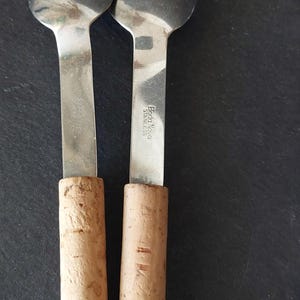 May include: Two stainless steel cheese knives with cork handles. The blades are round and the handles are cylindrical. The brand name "Eda Nova Stainless" is visible on the blade. The knives are on a dark surface.