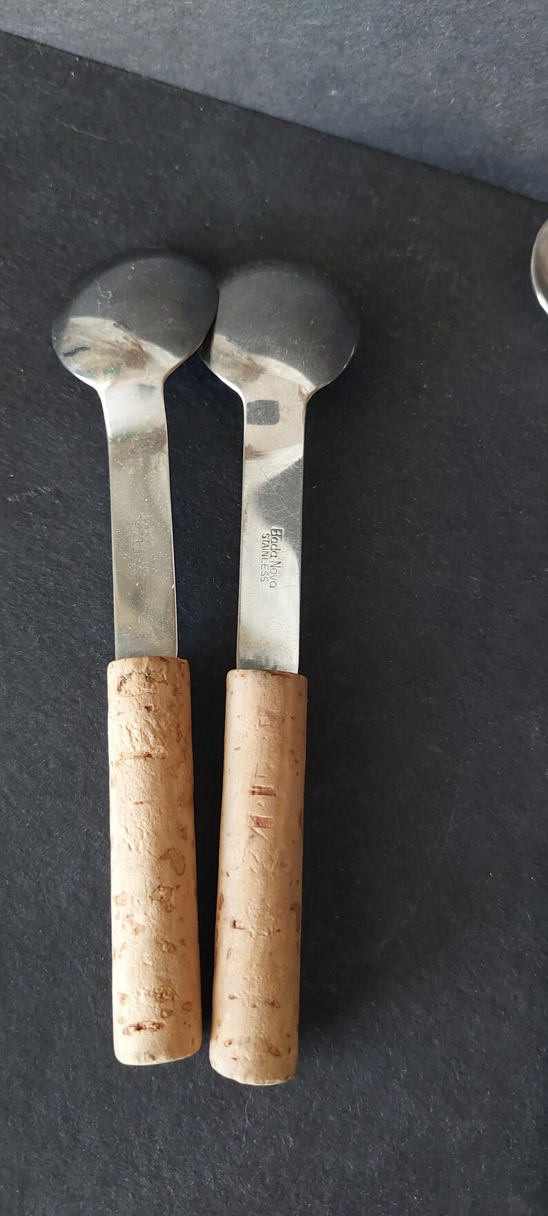 May include: Two stainless steel cheese knives with cork handles. The blades are round and the handles are cylindrical. The brand name "Eda Nova Stainless" is visible on the blade. The knives are on a dark surface.