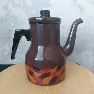 Puede incluir: Una cafetera de esmalte marrón con asa y tapa negras. La cafetera presenta un patrón decorativo de formas de llama naranja, roja y negra alrededor de la base. El pico se curva hacia arriba.