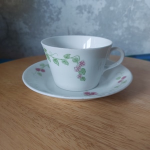 Puede incluir: Una taza de té y un platillo blancos con un diseño floral. La taza tiene una vid verde con flores rosas y hojas verdes. El platillo tiene un diseño similar con flores rosas.