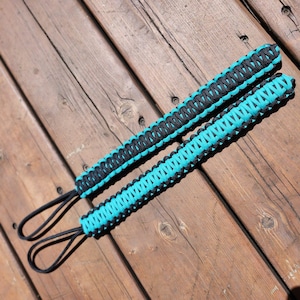 May include: Two paracord wrist straps, each featuring a black and turquoise woven design. The straps have a loop at the end for easy attachment. The items are displayed on a weathered wooden surface.