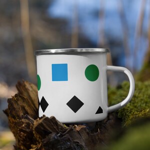 May include: A white enamel mug with a blue square, green circle, and black diamond shapes. The mug has a silver rim and handle.