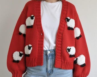 Könnte beinhalten: Ein leuchtend roter Cardigan mit Applikationen von weißen und schwarzen Schafen. Der Strickpullover hat einen Knopfverschluss und lange Ärmel. Die Person trägt ein weißes T-Shirt und blaue Jeans.