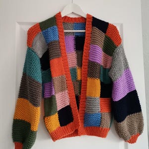 May include: A multi-coloured patchwork cardigan. The open-fronted sweater features squares of orange, green, blue, purple, and grey. It has long sleeves and is hanging on a white hanger against a white door.