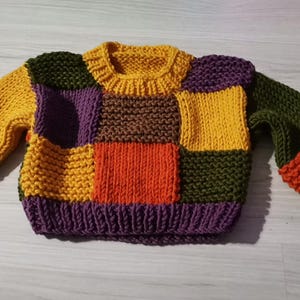 Op de afbeelding: Een kleurrijke patchwork trui in verschillende tinten geel, paars, oranje, groen en bruin. De gebreide trui heeft een geribbelde kraag, manchetten en zoom. De mouwen en het lijfje bestaan uit verschillende gekleurde vierkanten.