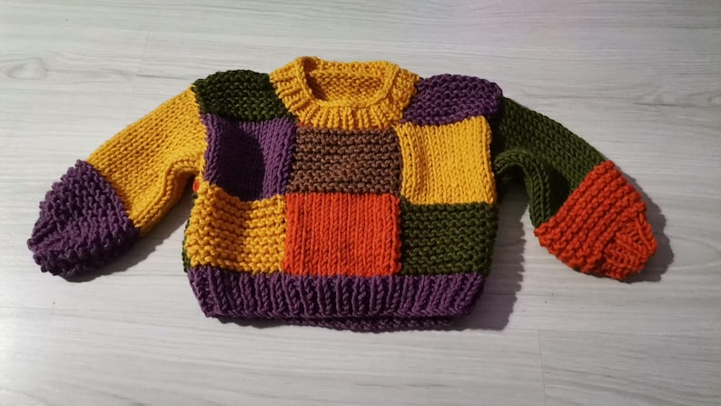 Op de afbeelding: Een kleurrijke patchwork trui in verschillende tinten geel, paars, oranje, groen en bruin. De gebreide trui heeft een geribbelde kraag, manchetten en zoom. De mouwen en het lijfje bestaan uit verschillende gekleurde vierkanten.