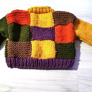 Op de afbeelding: Een kleurrijke patchwork-trui in verschillende kleuren, waaronder oranje, groen, geel, paars en bruin. De trui is gebreid met een geribbelde kraag en manchetten. De mouwen en de body bestaan uit vierkanten in verschillende kleuren.