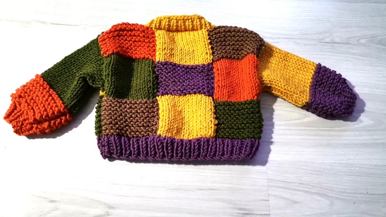 Op de afbeelding: Een kleurrijke patchwork-trui in verschillende kleuren, waaronder oranje, groen, geel, paars en bruin. De trui is gebreid met een geribbelde kraag en manchetten. De mouwen en de body bestaan uit vierkanten in verschillende kleuren.
