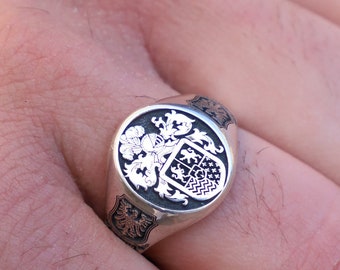 May include: A silver signet ring with an intricate design featuring a coat of arms. The ring is oval shaped and has a black enamel background.