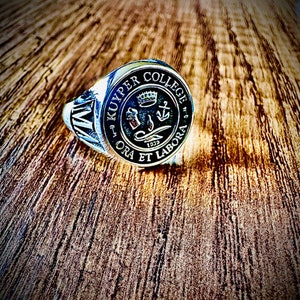 Puede incluir: Un anillo de plata con un escudo negro y dorado con las palabras "Kuypers College" y "Ora Et Labora" y el año 1939.