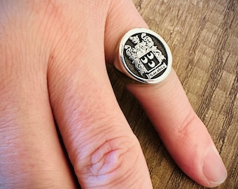 May include: A silver signet ring with a black enamel crest featuring a coat of arms and the text "Sarabia".